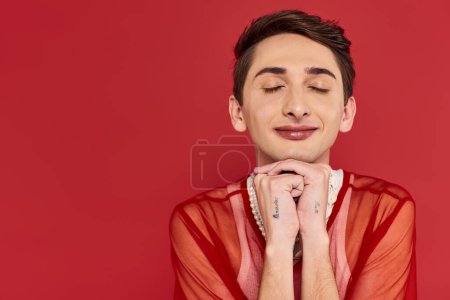 Téléchargez les photos : Séduisant homme androgyne modish en tenue décontractée avec résille rouge vibrant posant les yeux fermés - en image libre de droit
