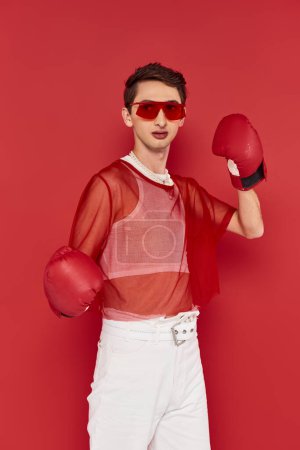 fashionable androgynous man with red fishnet posing actively with boxing gloves and looking away