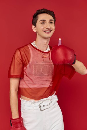 Téléchargez les photos : Gai homme androgyne à la mode avec des gants de boxe tenant rouge à lèvres et souriant à la caméra - en image libre de droit