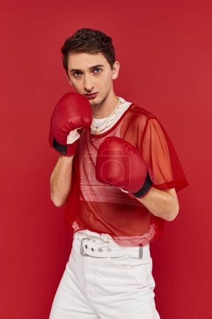 Téléchargez les photos : Bel homme gay bien habillé en tenue décontractée avec des gants de boxe sur fond rouge regardant la caméra - en image libre de droit