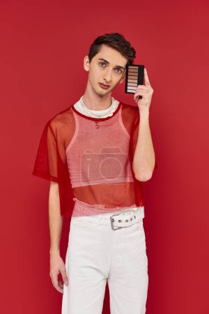 Téléchargez les photos : Bel homme androgyne avec résille rouge tenant la palette des ombres à paupières et regardant la caméra - en image libre de droit