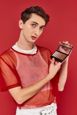 Photo for Handsome androgynous man with red fishnet holding eye shadows palette and looking at camera - Royalty Free Image