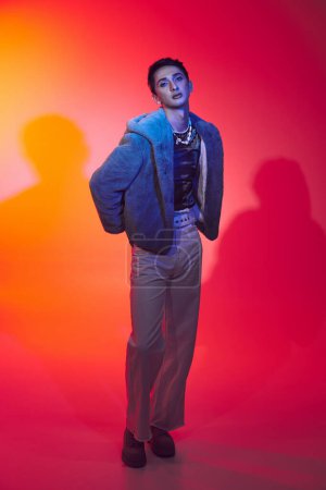 fashionable androgynous man in purple faux fur posing on vibrant backdrop and looking at camera