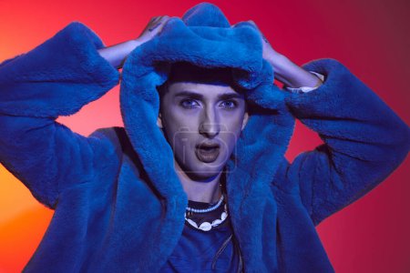 fashionable androgynous man in purple faux fur posing on vibrant backdrop and looking at camera