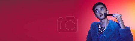 Photo for Elegant androgynous man in stylish attire using makeup brush and posing on vibrant backdrop, banner - Royalty Free Image