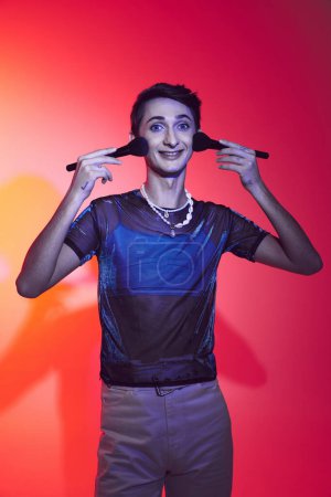 Photo for Cheerful elegant androgynous man in casual attire holding two makeup brushes and looking at camera - Royalty Free Image