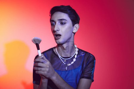 Photo for Sophisticated androgynous man in stylish attire using makeup brush and posing on vibrant backdrop - Royalty Free Image