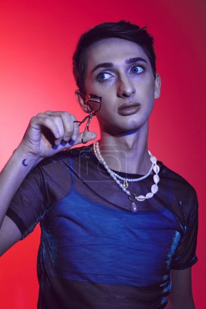 handsome fashionable androgynous man in casual attire holding eyelash curler on vibrant backdrop