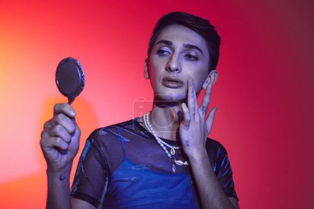 Photo for Alluring androgynous man in vivid attire looking at small mirror while on red and orange backdrop - Royalty Free Image