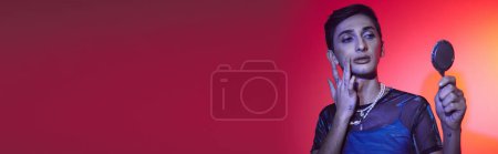 Photo for Handsome androgynous man in casual attire looking at mirror while on red and orange backdrop, banner - Royalty Free Image