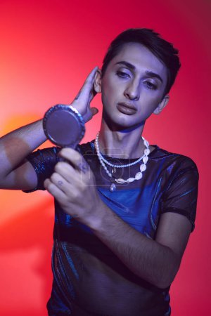 Téléchargez les photos : Alluring androgynous man in vivid attire looking at small mirror while on red and orange backdrop - en image libre de droit