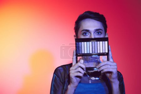Photo for Enticing gay man in casual attire using eyeshadows palette and looking at camera on vibrant backdrop - Royalty Free Image