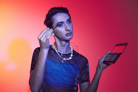 Téléchargez les photos : Voguish gay l'homme en tenue décontractée avec accessoires en utilisant fard à paupières palette sur fond vibrant - en image libre de droit