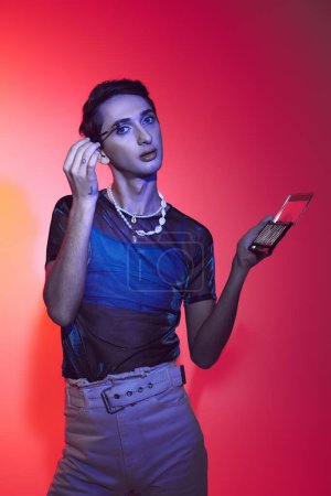 Téléchargez les photos : Enticing gay man in casual attire using eyeshadows palette and looking at camera on vibrant backdrop - en image libre de droit
