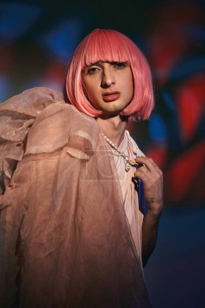 Foto de Atractivo de moda gay hombre usando elegante rosa peluca y mirando la cámara en colorido fondo - Imagen libre de derechos