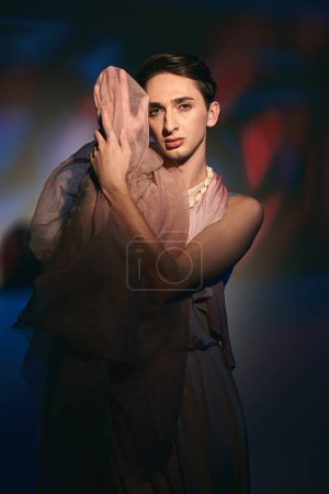 enticing androgynous fashionista with dark hair in pastel attire looking at camera on vivid backdrop