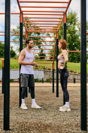 A determined woman, guided by a personal trainer, engage in outdoor exercise beneath a metal structure.