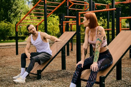 A man and a woman in sportswear sit on benches, exercising together in a park. Their determination and motivation are evident.