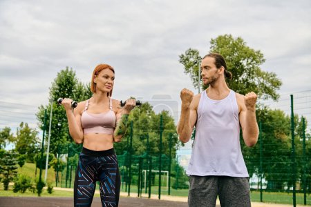 A determined man and woman in sportswear stand together outdoors, exercising with dumbbells