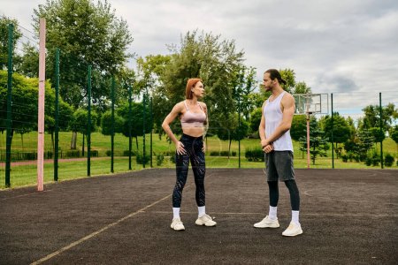 Un hombre y una mujer, usando ropa deportiva, se paran en una cancha de baloncesto, mostrando determinación y motivación durante su sesión de ejercicio al aire libre.