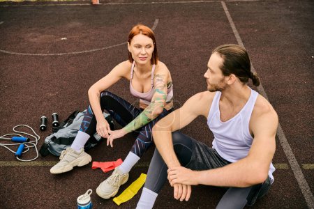 A man and woman in sportswear, resting on a basketball court, exuding determination and motivation.
