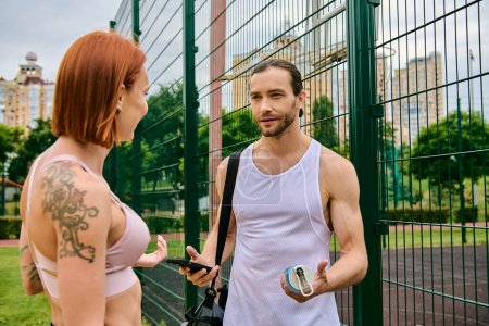 A man and a woman in sportswear stand by a fence, motivated by personal training in an outdoor workout session.