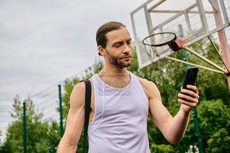 A man holds a cell phone in front of a basketball hoop.