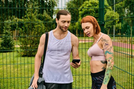 A determined man and woman in sportswear stand in front of a fence, using smartphone