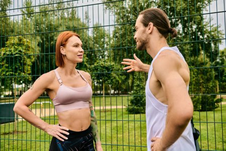 A determined woman talks with a personal trainer while exercising in front of a fence outdoors.