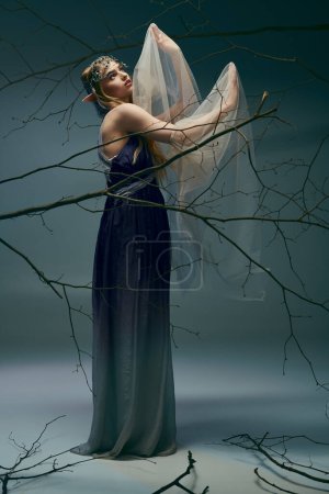 Téléchargez les photos : Une jeune femme en robe longue tient délicatement un voile, exsudant une aura de fée dans un décor de studio. - en image libre de droit