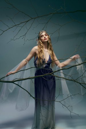 A young woman in a blue dress stands gracefully in front of a sprawling tree branch in a whimsical studio setting.