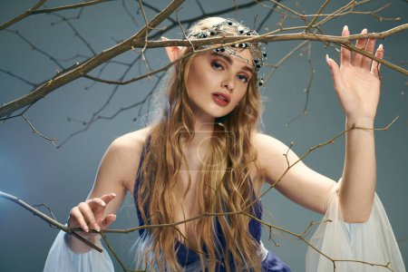 Una joven con un vestido azul parecido a una princesa elfa sostiene delicadamente una rama floreciente en un ambiente de estudio.