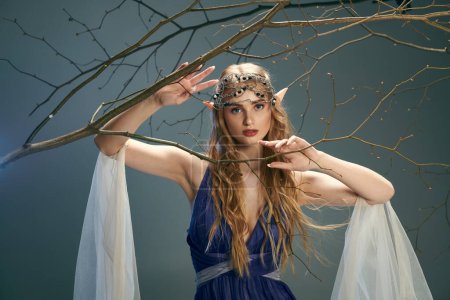 A young woman in a blue dress resembling an elf princess delicately holds a branch in a whimsical, fairy-like studio setting.