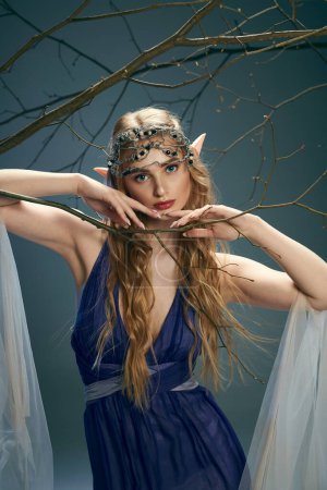 Une jeune femme en robe bleue tient une branche délicate dans un décor de studio, incarnant un personnage de conte de fées.