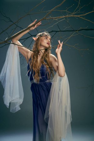 Une jeune femme en robe bleue ressemblant à une princesse elfe, tient délicatement une branche dans un décor de studio.