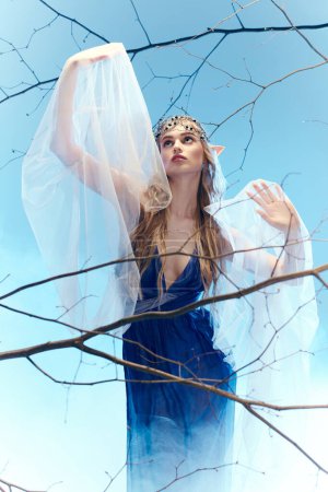 A young woman in a blue dress gracefully holds a white veil, exuding a fairy-like aura in a mystical studio setting.