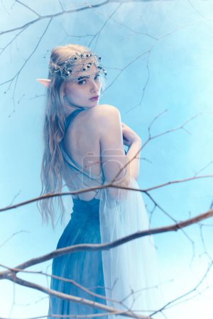 Téléchargez les photos : Une jeune femme dans une robe bleue fluide se tient gracieusement dans un arbre, incarnant une princesse fée dans un monde fantastique. - en image libre de droit