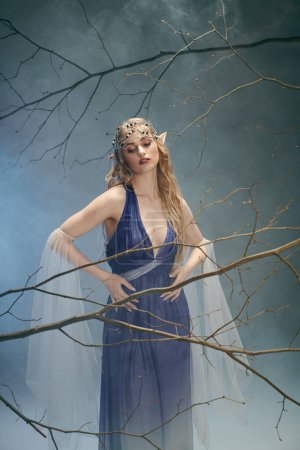 Une jeune femme en robe bleue se tient gracieusement devant un arbre majestueux, incarnant une présence féerique dans un décor de studio.