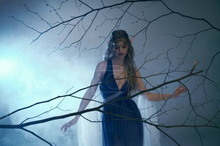 A young woman with elf princess vibes stands gracefully in front of a tree, wearing a stunning blue dress.