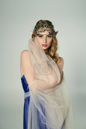 Eine junge Frau in einem blauen Kleid mit Schleier auf dem Kopf, verkörpert das Wesen einer mystischen Elfenprinzessin in einem skurrilen Studio-Setting.