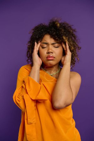 Photo for Emotional African American woman in orange shirt holds head in distress. - Royalty Free Image