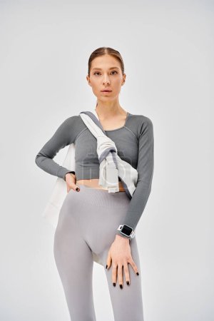 A sporty young woman exudes elegance in her gray top and leggings against a grey background.