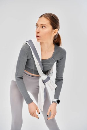 A sporty young woman stands confidently with a white scarf gracefully draped around her neck against a grey background.