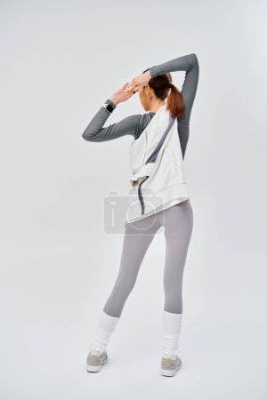 A sporty young woman in grey leggings and a white top poses gracefully against a grey background.