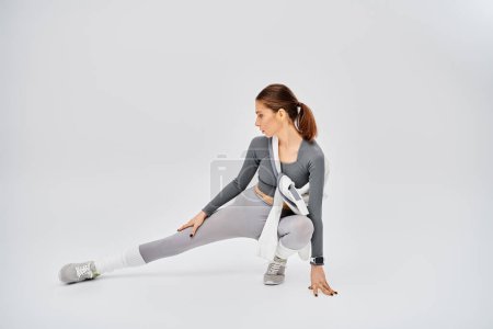 Jeune femme sportive en tenue active étire gracieusement son corps sur un fond gris.