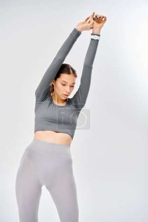 Una joven deportista en ropa deportiva gris logra una pose de yoga con fuerza y equilibrio sobre un fondo gris.