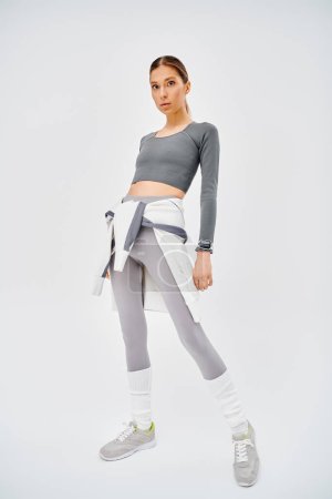A sporty young woman in grey and white attire striking a confident pose against a grey background.