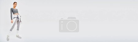 Téléchargez les photos : Une jeune femme sportive dans un haut gris et pantalon blanc frappe une pose gracieuse sur un fond gris. - en image libre de droit