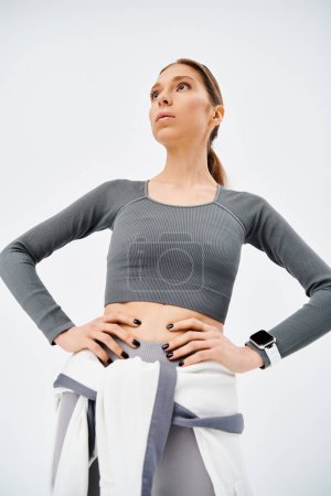 Una joven deportista en ropa activa se para con confianza con las manos en las caderas sobre un fondo gris.