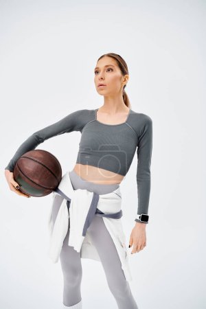 Eine sportliche junge Frau in aktiver Kleidung hält vor grauem Hintergrund selbstbewusst einen Basketball in der rechten Hand.